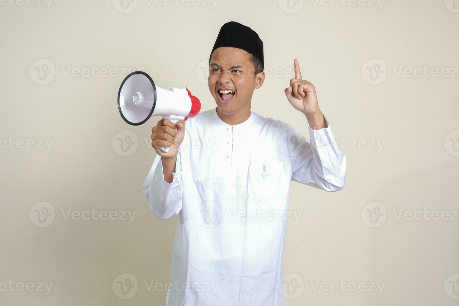 Portrait of attractive Asian muslim man in white shirt with skullcap speaking louder using megaphone, promoting product. Advertising concept. Isolated image on grey background photo