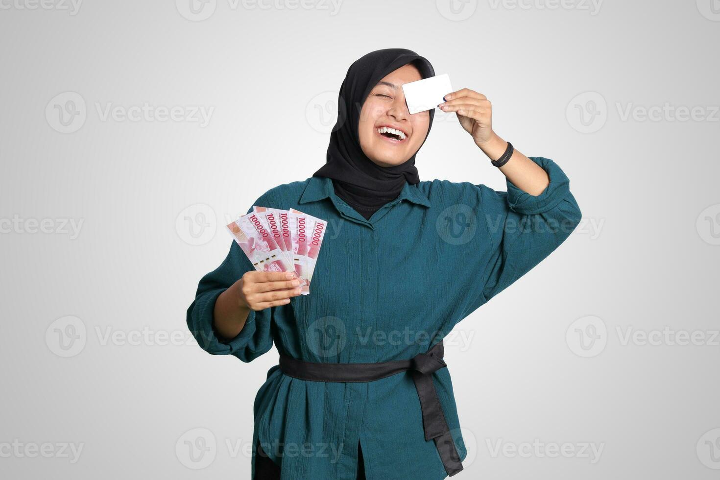 Portrait of cheerful Asian muslim woman with hijab, showing one hundred thousand rupiah while holding a credit card. Financial and savings concept. Isolated image on white background photo