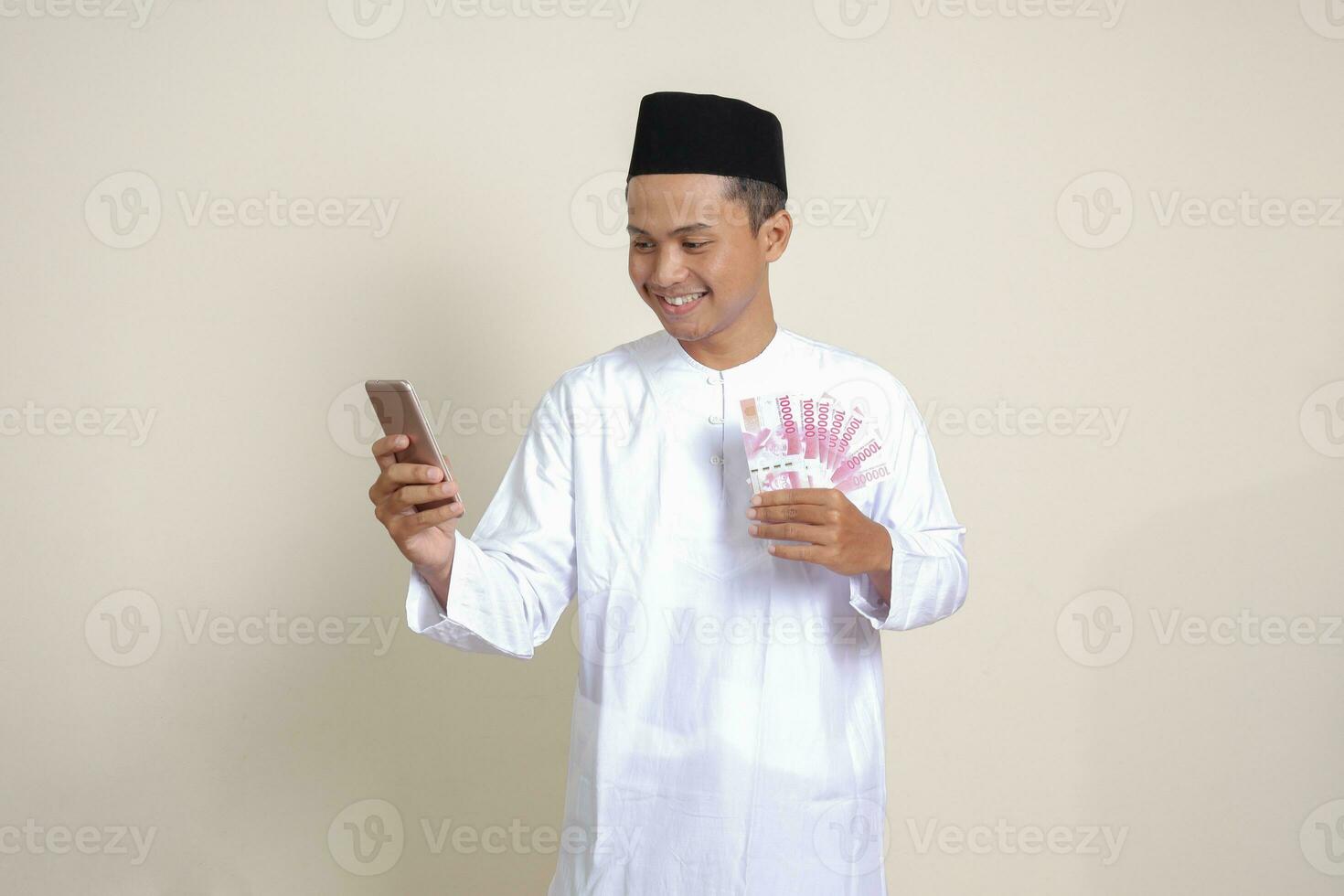 Portrait of attractive Asian muslim man in white shirt showing one hundred thousand rupiah while using mobile phone. Financial and savings concept. Isolated image on gray background photo