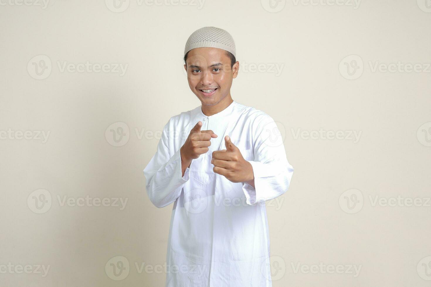 retrato de atractivo asiático musulmán hombre en blanco camisa con casquete demostración producto y señalando con su mano y dedo a el lado. publicidad concepto. aislado imagen en gris antecedentes foto