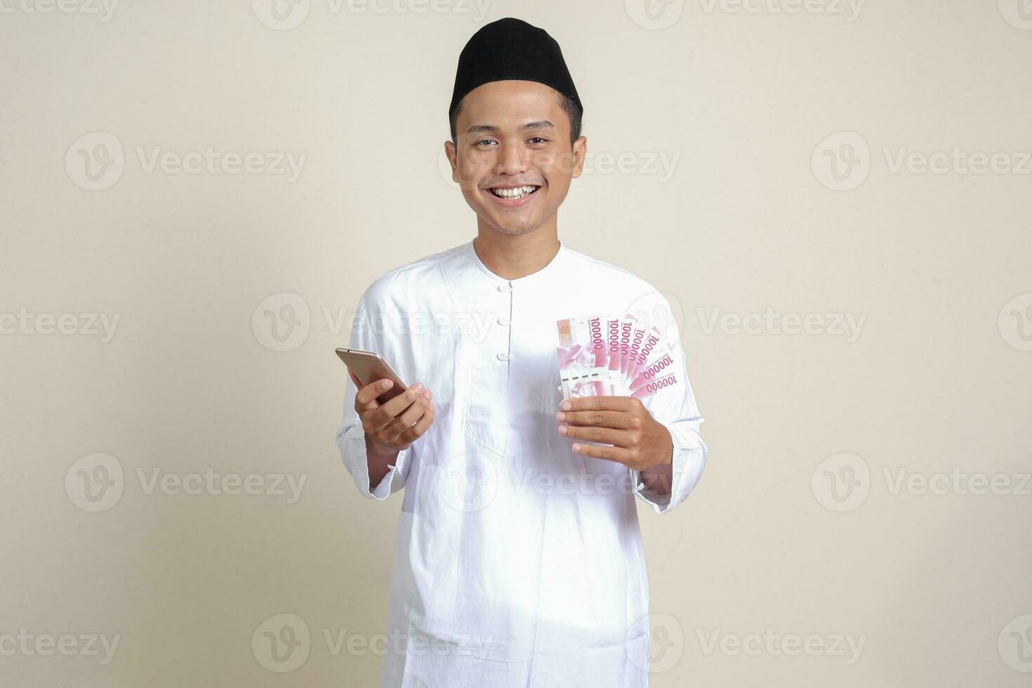 Portrait of attractive Asian muslim man in white shirt showing one hundred thousand rupiah while using mobile phone. Financial and savings concept. Isolated image on gray background photo