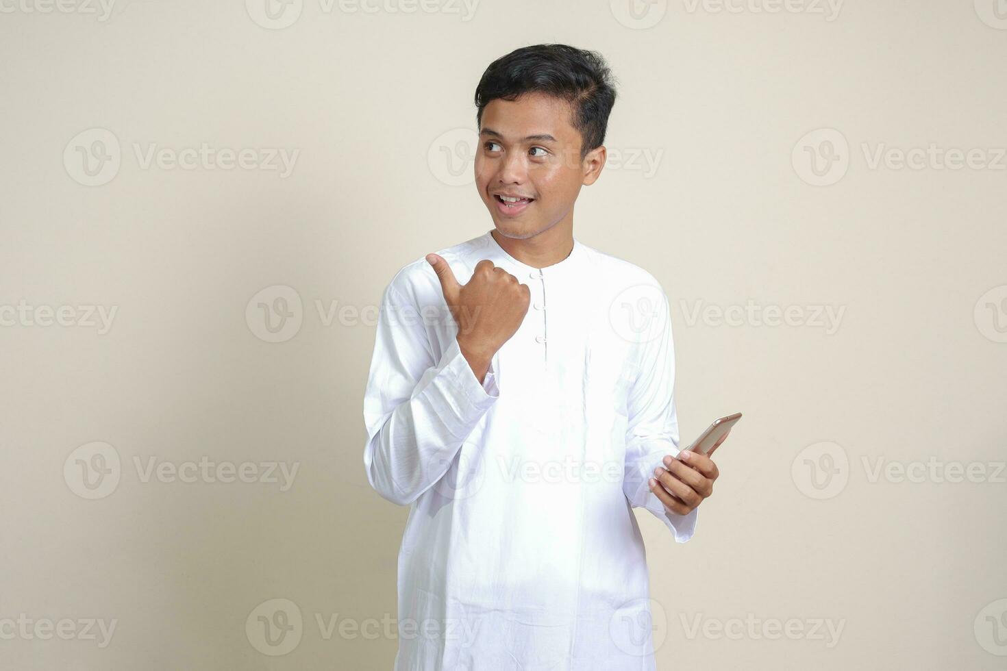 Portrait of attractive Asian muslim man in white shirt holding mobile phone with smiling expression on face while pointing finger to the side. Advertising concept. Isolated image on gray background photo