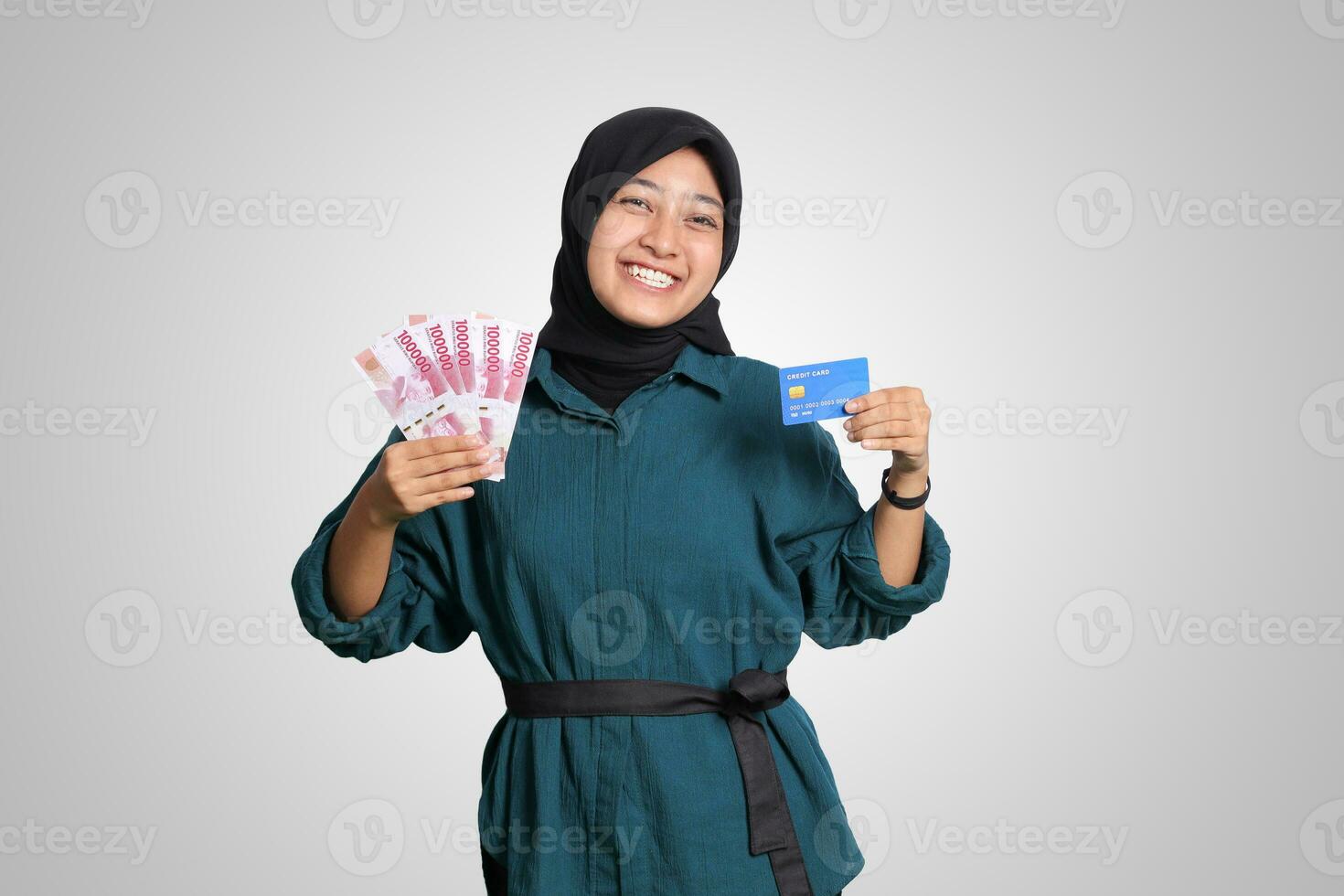 Portrait of cheerful Asian muslim woman with hijab, showing one hundred thousand rupiah while holding a credit card. Financial and savings concept. Isolated image on white background photo