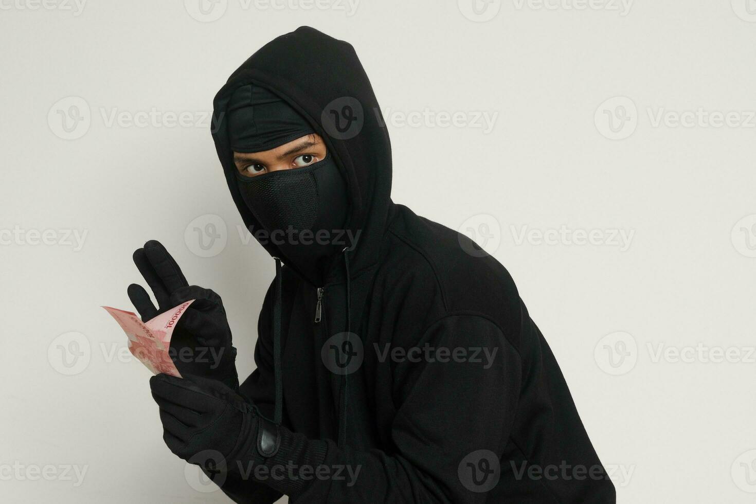 Portrait of mysterious man wearing black hoodie and mask stealing one hundred thousand rupiah successfully from victim. Isolated image on gray background photo