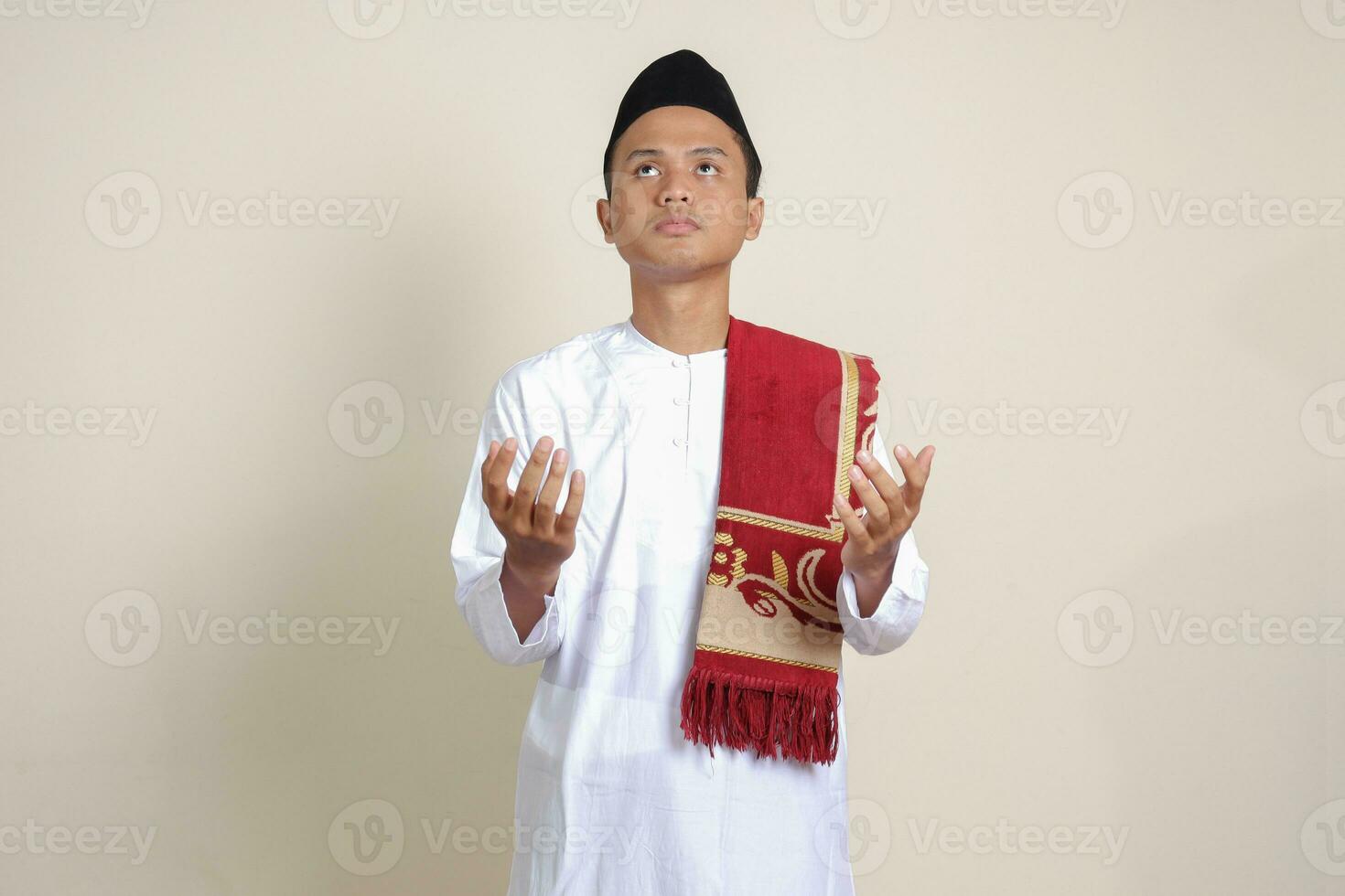 Portrait of attractive Asian muslim man in white shirt with skullcap praying earnestly with his hands raised. Isolated image on gray background photo