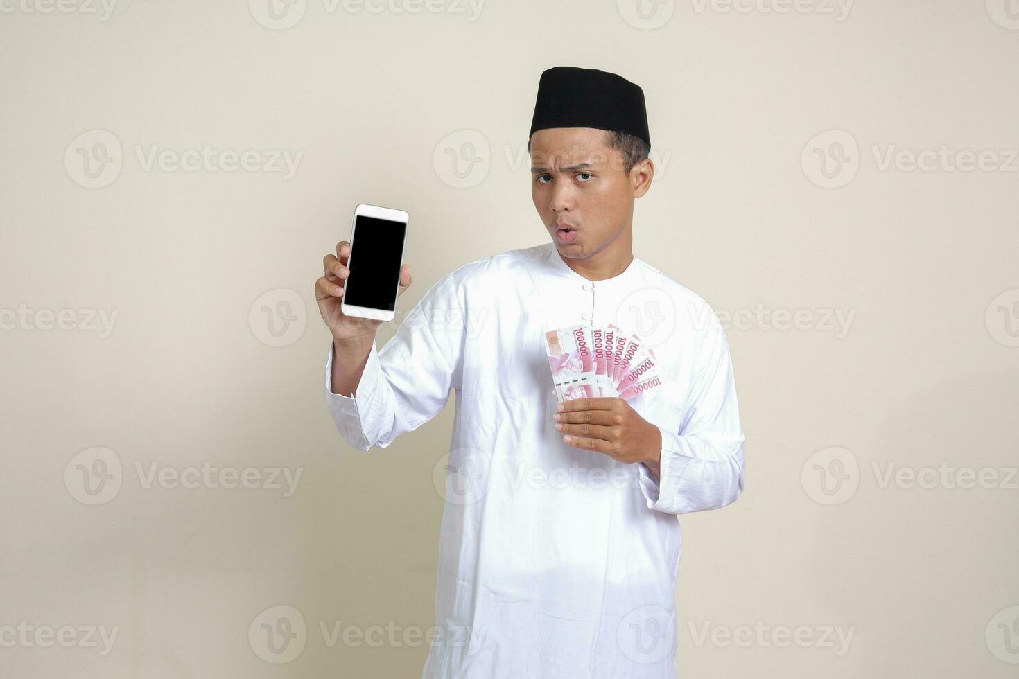 retrato de atractivo asiático musulmán hombre en blanco camisa con casquete demostración uno cien mil rupia mientras demostración blanco pantalla móvil teléfono. financiero y compras concepto. aislado imagen en gris foto