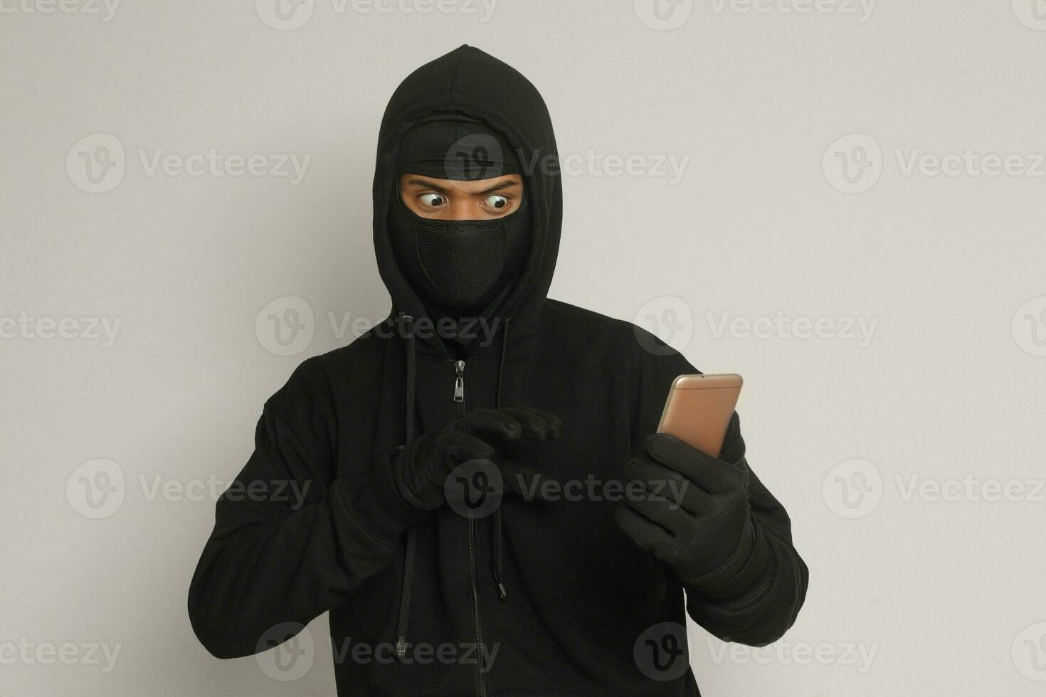 Portrait of mysterious man wearing black hoodie and mask doing hacking activity on mobile phone, hacker holding a smartphone. Isolated image on gray background photo