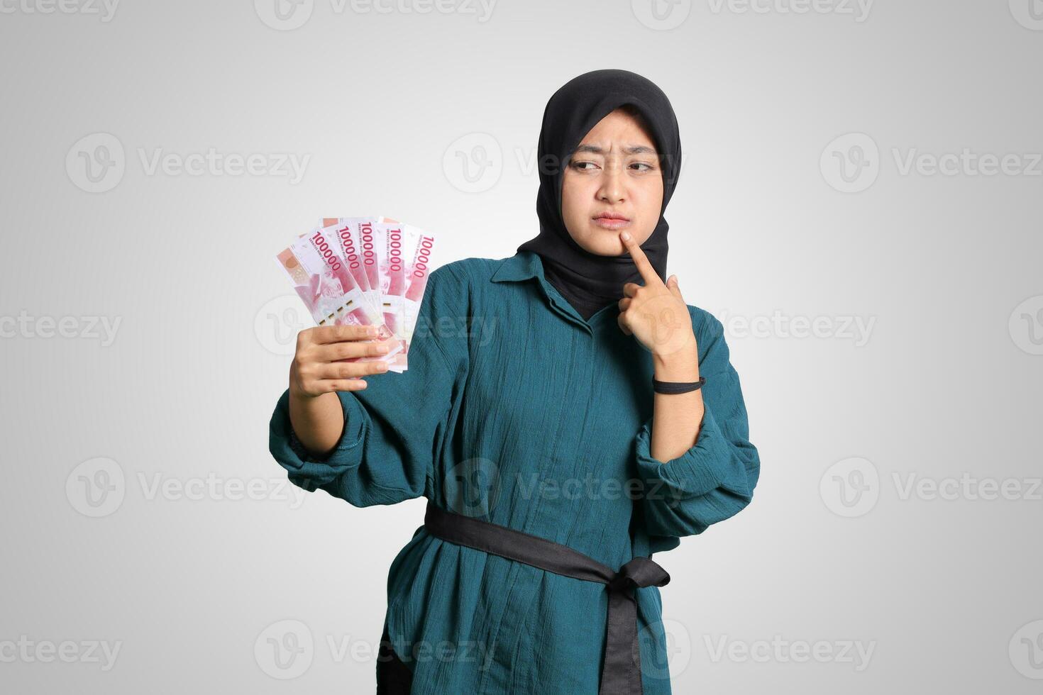 Portrait of confused Asian hijab woman in casual outfit showing one hundred thousand rupiah while thinking with hand on chin. Financial and savings concept. Isolated image on white background photo