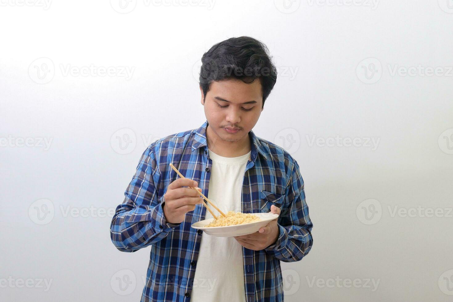 retrato de contento asiático hombre en azul tartán camisa comiendo delicioso instante tallarines con palillos servido en lámina. aislado imagen en blanco antecedentes foto