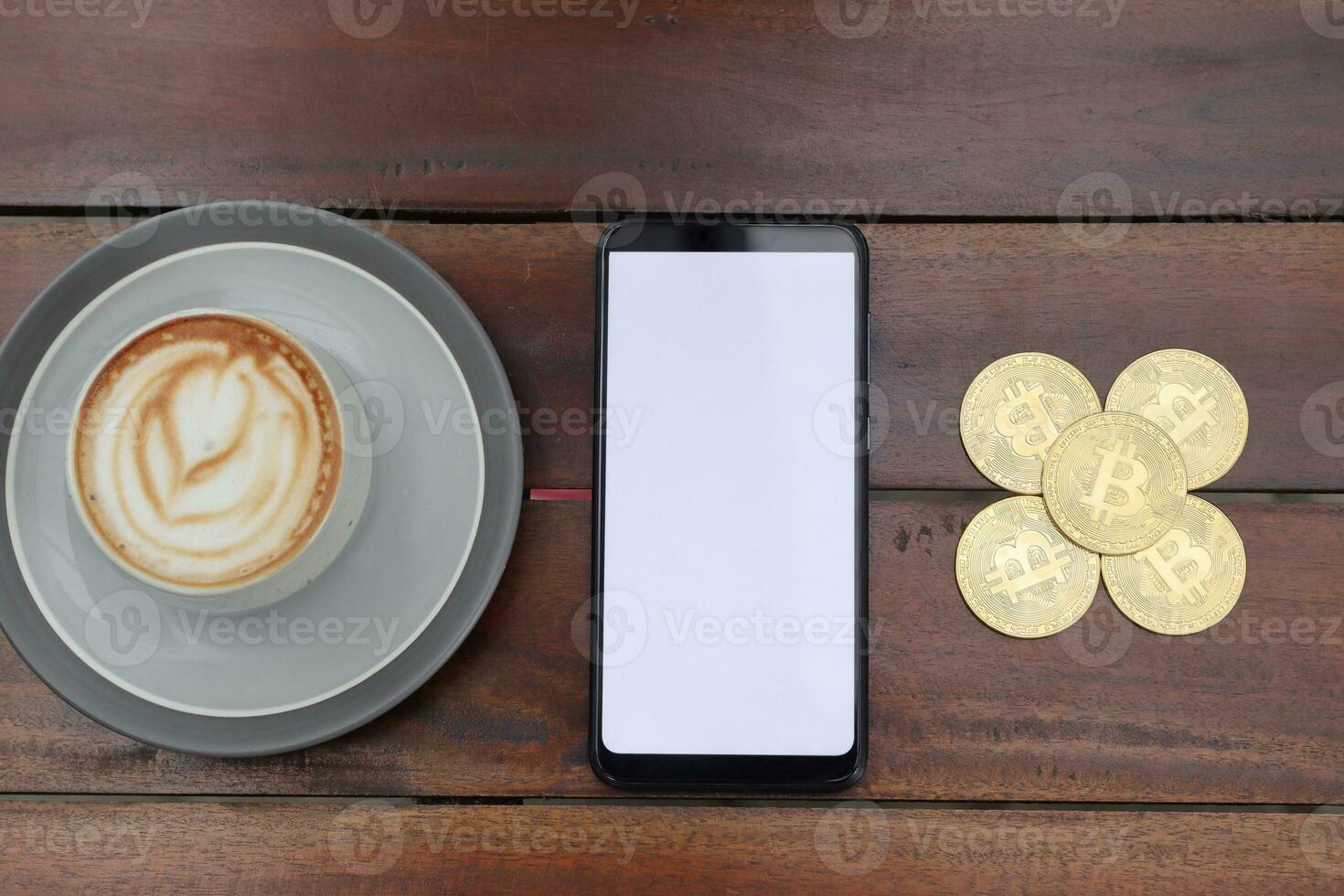 Top view of blank screen mobile phone with a pile of bitcoin and cup of coffee on wooden table. Cryptocurrency trading concept photo