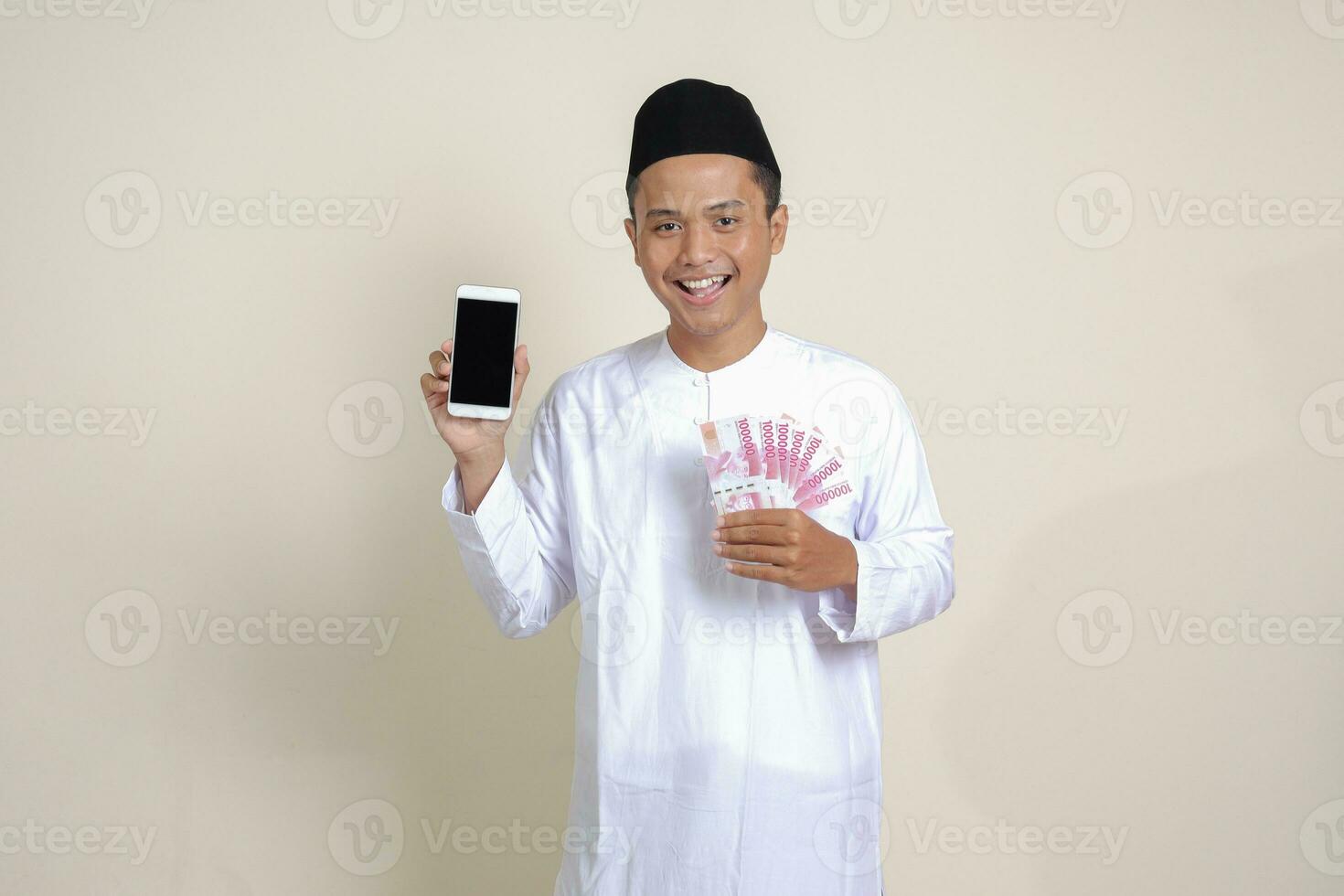 Portrait of attractive Asian muslim man in white shirt with skullcap showing one hundred thousand rupiah while showing blank screen mobile phone. Financial and shopping concept. Isolated image on gray photo