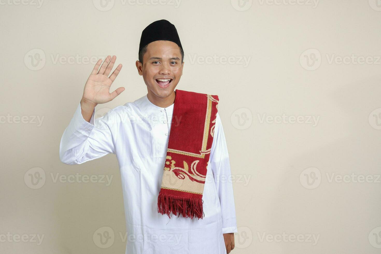 Portrait of attractive Asian muslim man in white shirt saying hi, waving hand at camera, greeting person with happy smile. Isolated image on gray background photo