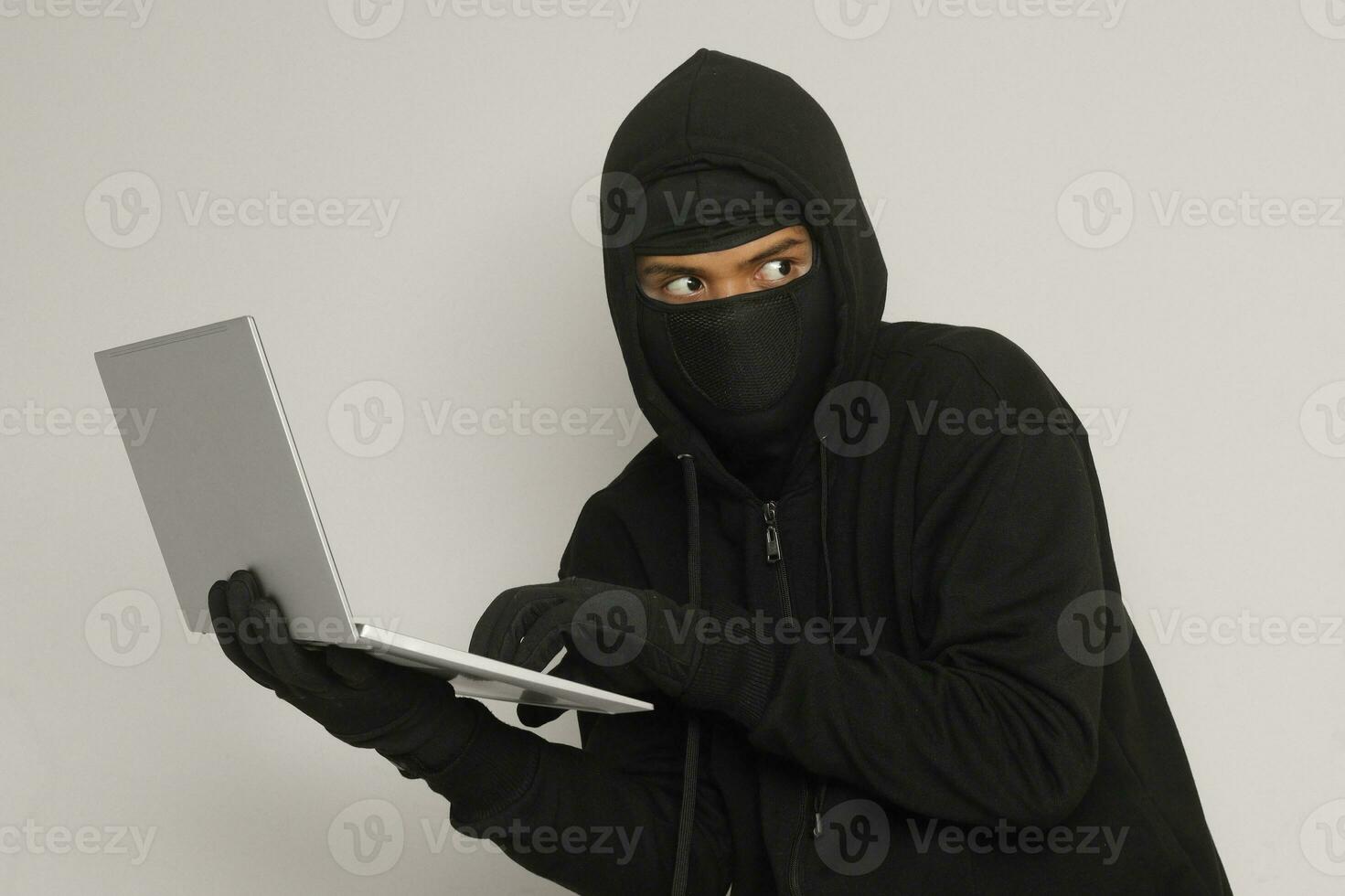 Portrait of mysterious man wearing black hoodie and mask doing hacking activity on laptop, hacker holding a personal computer. Cyber security concept. Isolated image on gray background photo