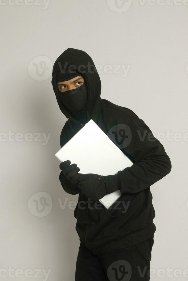 Mysterious robber thief man wearing black hoodie and mask stealing laptop and sneak out. Isolated image on gray background photo