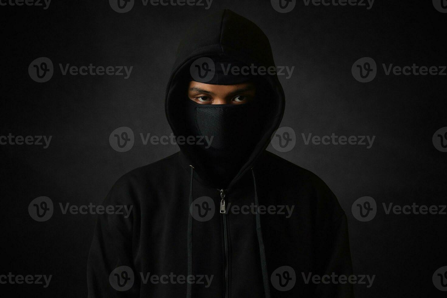 Mysterious man wearing black hoodie and mask standing against dark background, looking at camera. Dramatic low light portrait photo