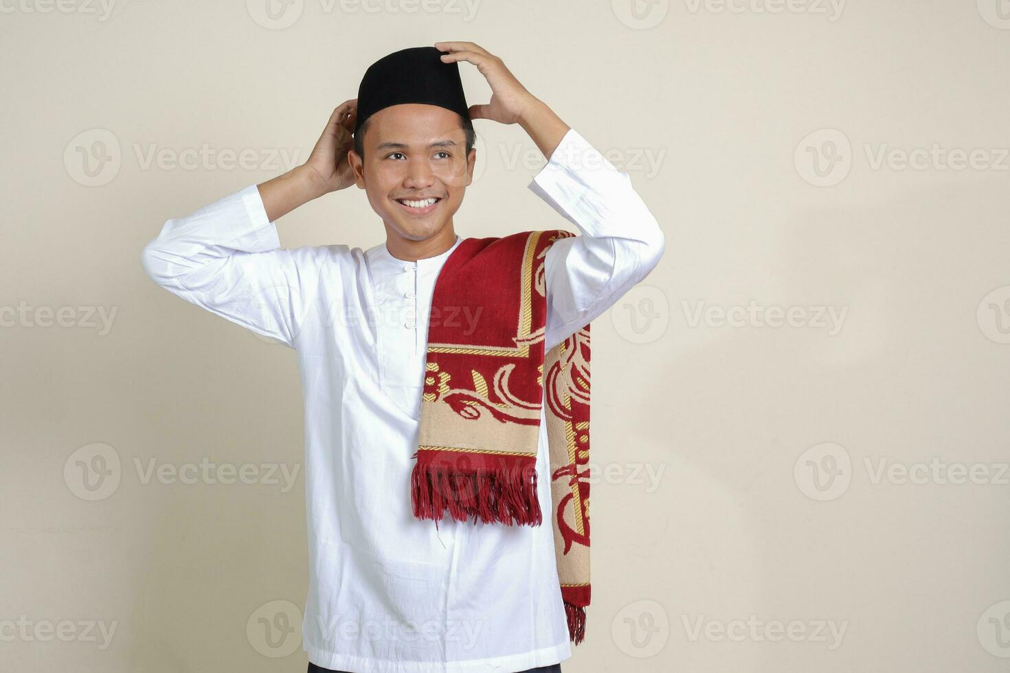 Portrait of attractive Asian muslim man in white shirt with skullcap trying to adjust his songkok or black skullcap. Isolated image on gray background photo