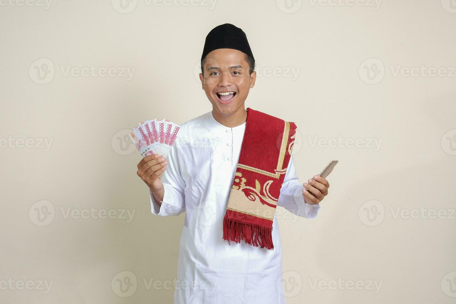 Portrait of attractive Asian muslim man in white shirt showing one hundred thousand rupiah while using mobile phone. Financial and savings concept. Isolated image on gray background photo