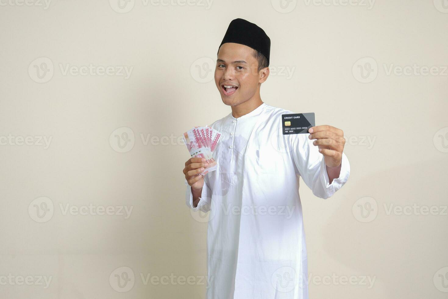 retrato de atractivo asiático musulmán hombre en blanco camisa con casquete participación uno cien mil rupia y presentación crédito tarjeta. aislado imagen en gris antecedentes foto