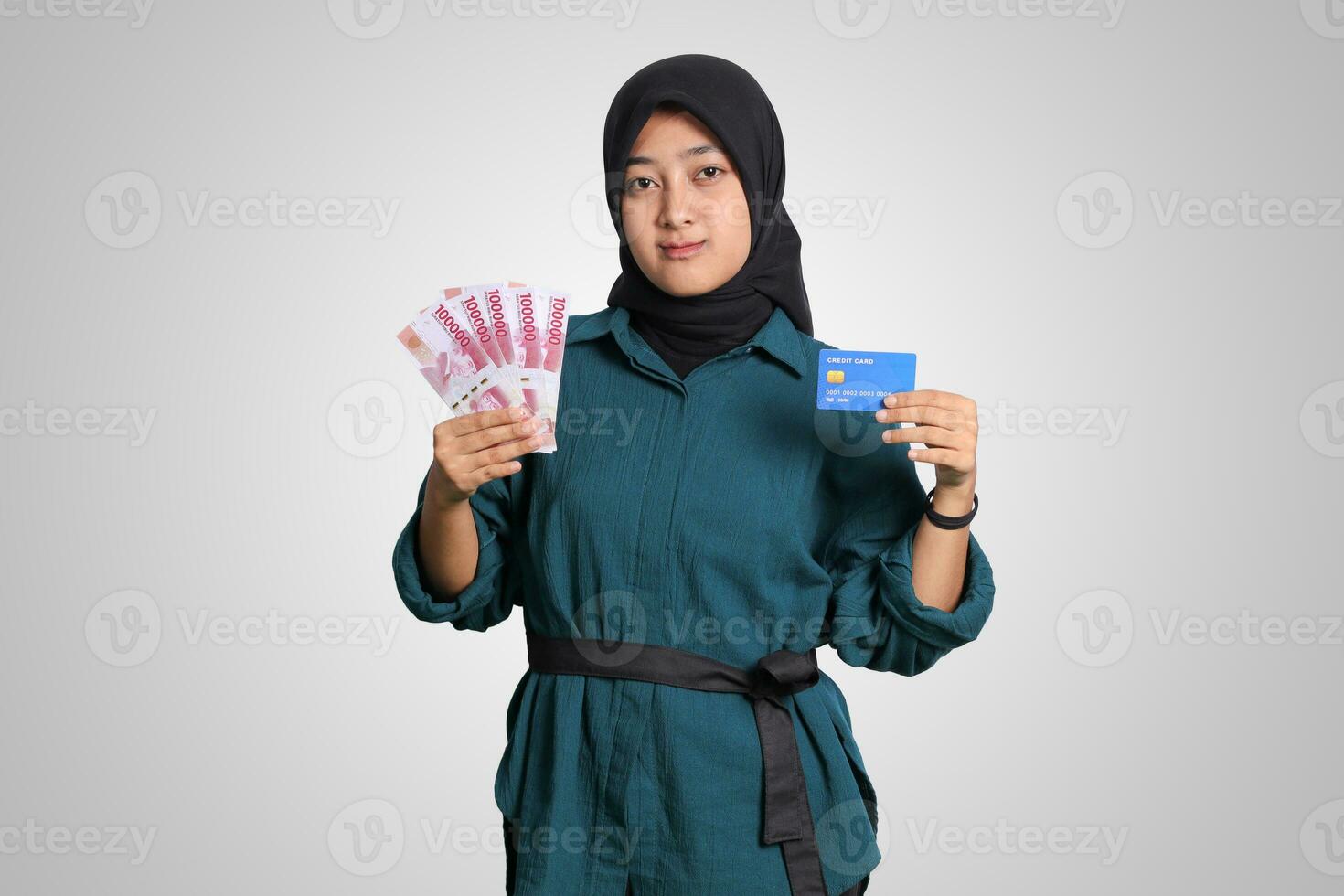 Portrait of cheerful Asian muslim woman with hijab, showing one hundred thousand rupiah while holding a credit card. Financial and savings concept. Isolated image on white background photo