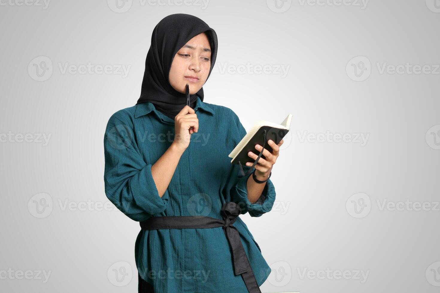 retrato de emocionado asiático musulmán mujer con hijab escritura en Nota libro y pensando con bolígrafo en mentón. publicidad concepto. aislado imagen en blanco antecedentes foto