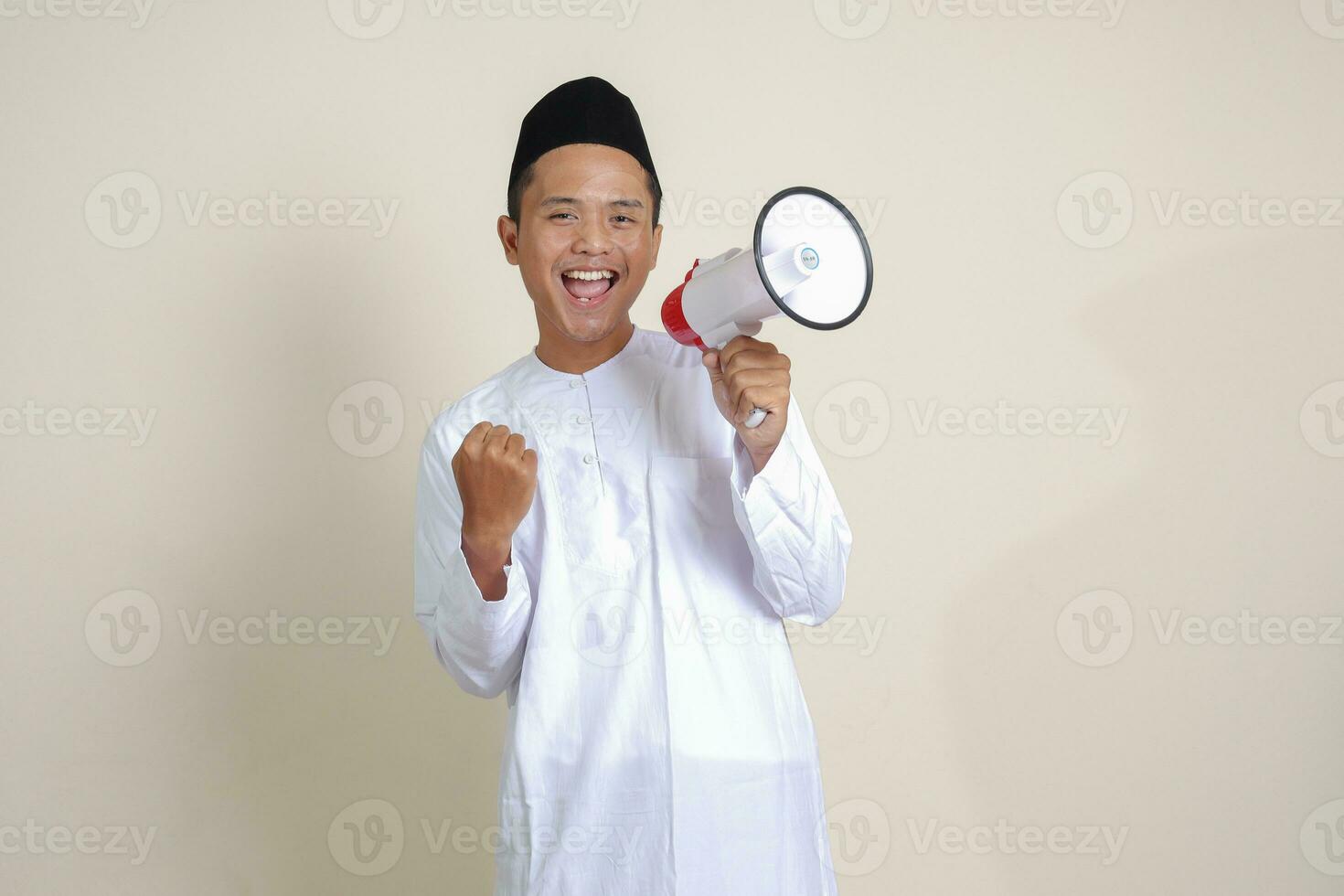 Portrait of attractive Asian muslim man in white shirt with skullcap speaking louder using megaphone, promoting product. Advertising concept. Isolated image on grey background photo