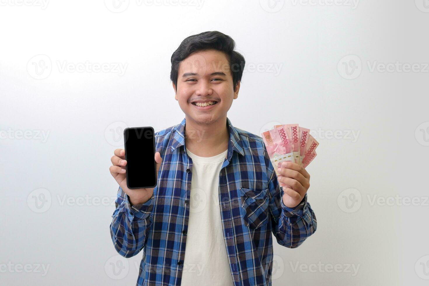 Portrait of excited Asian man in blue plaid shirt standing against white background, showing one hundred thousand rupiah while showing blank screen mobile phone. Financial and shopping concept. photo