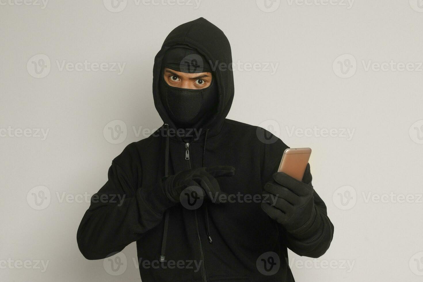 Portrait of mysterious man wearing black hoodie and mask doing hacking activity on mobile phone, hacker holding a smartphone. Isolated image on gray background photo