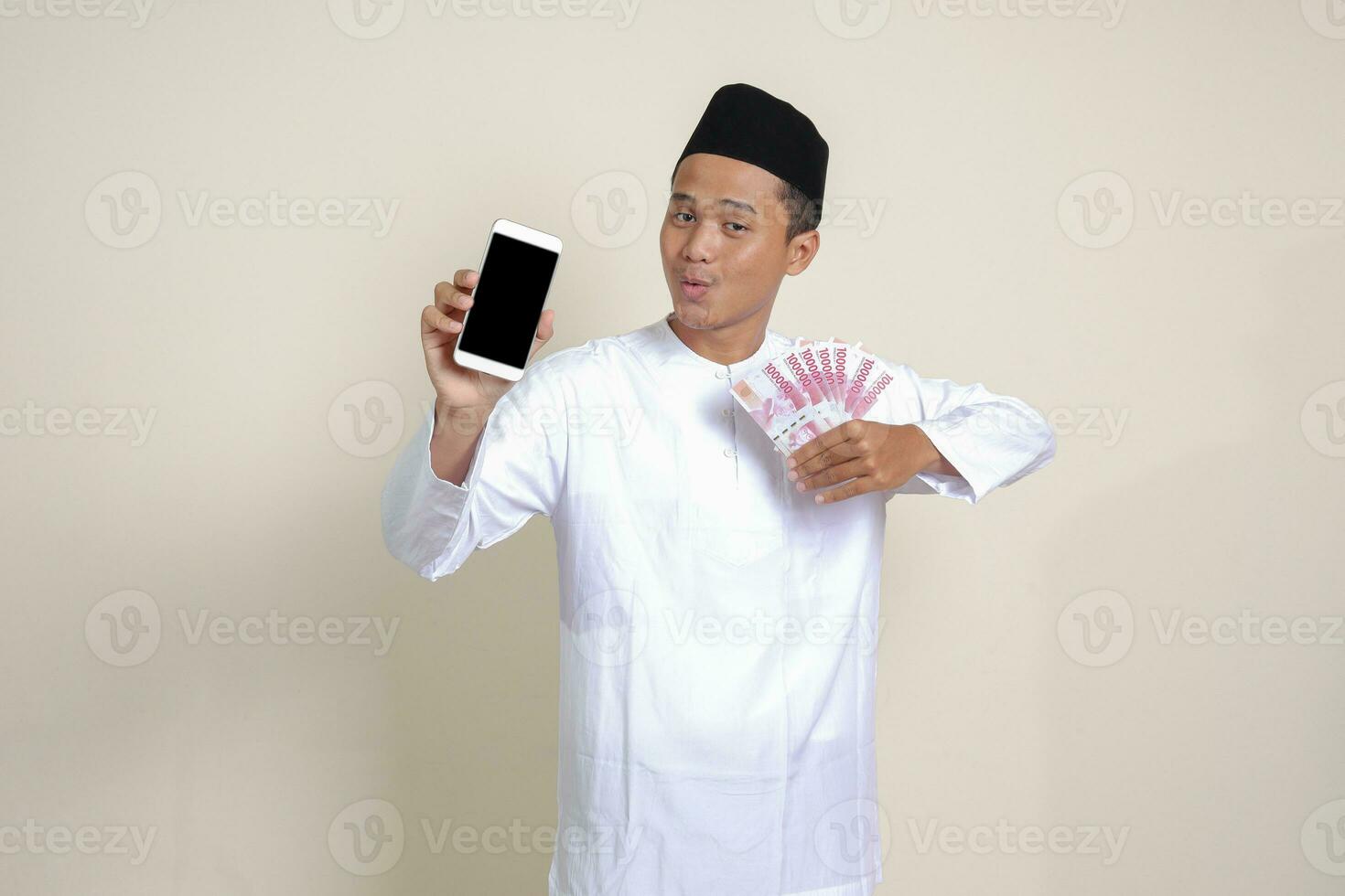 Portrait of attractive Asian muslim man in white shirt with skullcap showing one hundred thousand rupiah while showing blank screen mobile phone. Financial and shopping concept. Isolated image on gray photo