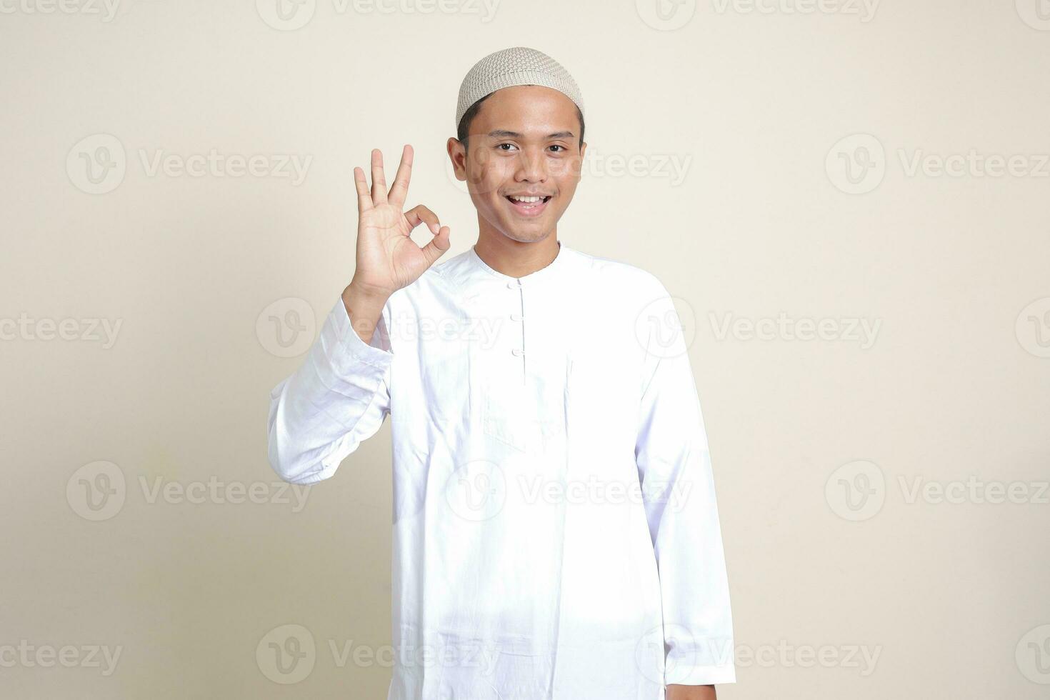 Portrait of attractive Asian muslim man in white shirt with skullcap showing ok hand gesture and smiling looking at camera. Advertising concept. Isolated image on gray background photo