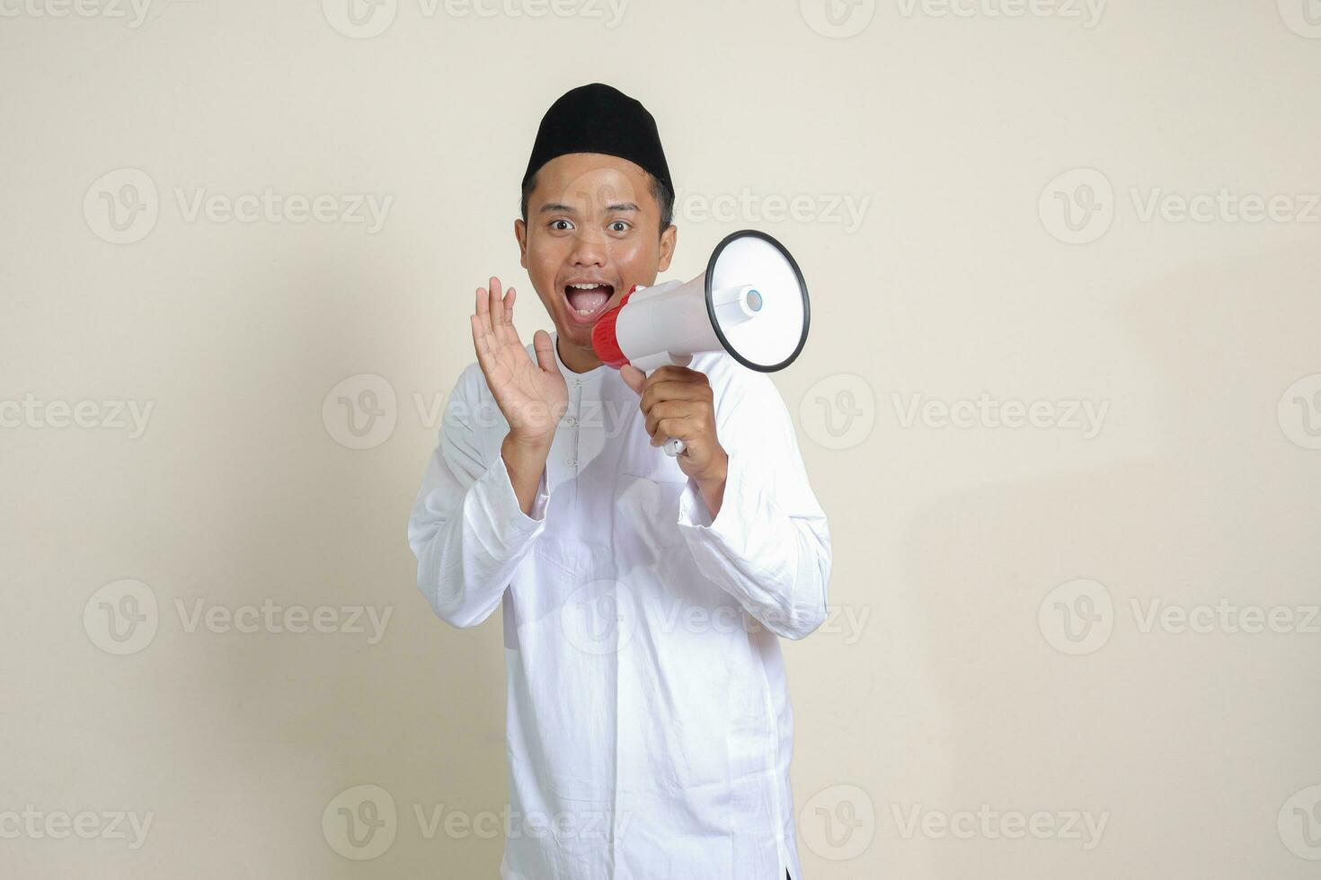 Portrait of attractive Asian muslim man in white shirt with skullcap speaking louder using megaphone, promoting product. Advertising concept. Isolated image on grey background photo