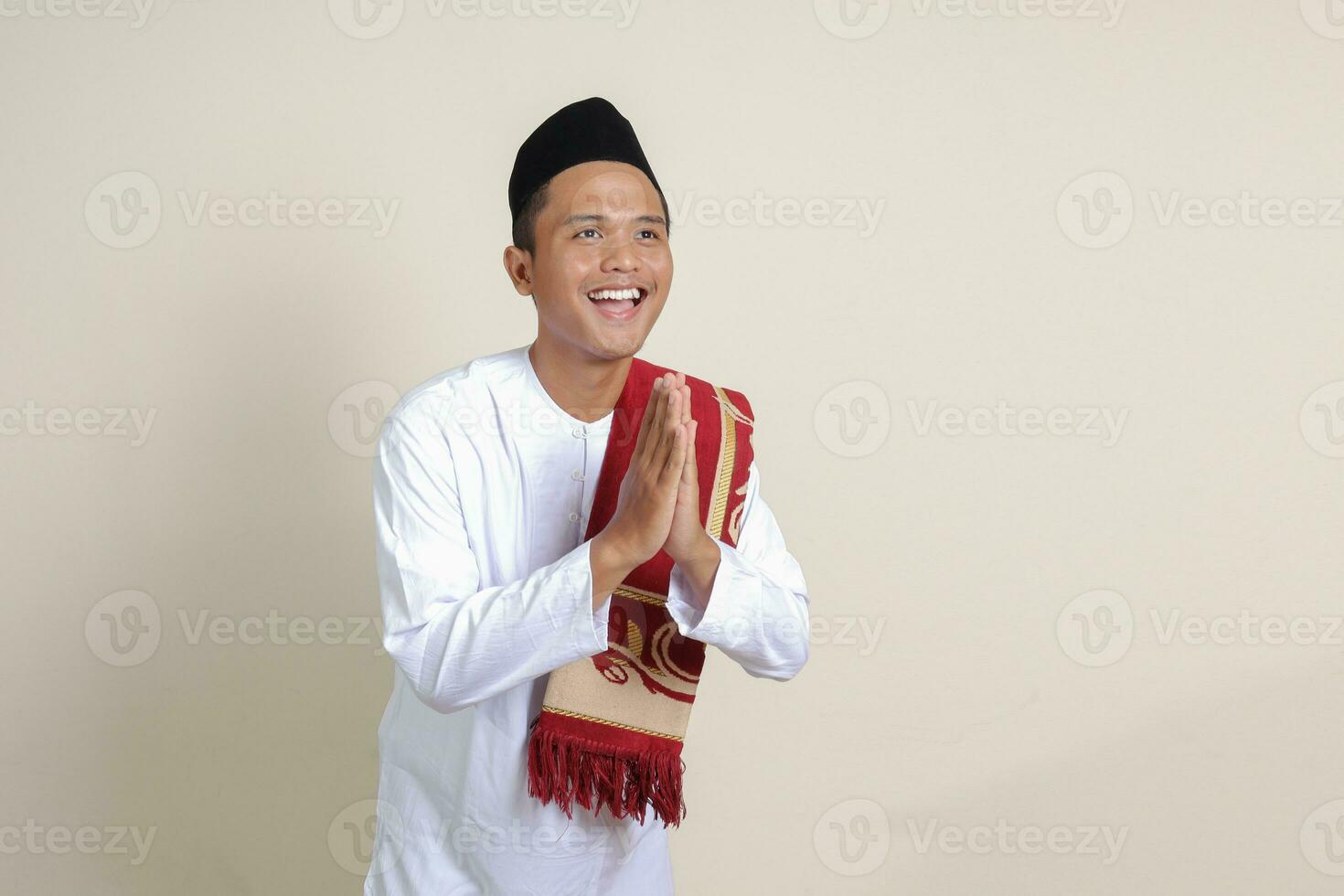 Portrait of attractive Asian muslim man in white shirt with skullcap showing apologize and welcome hand gesture. Isolated image on gray background photo