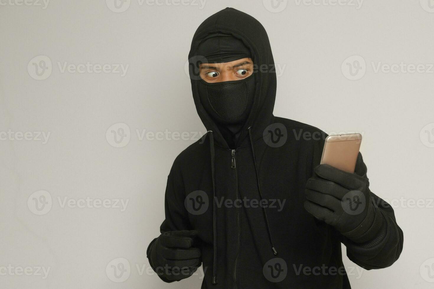 Portrait of mysterious man wearing black hoodie and mask doing hacking activity on mobile phone, hacker holding a smartphone. Isolated image on gray background photo