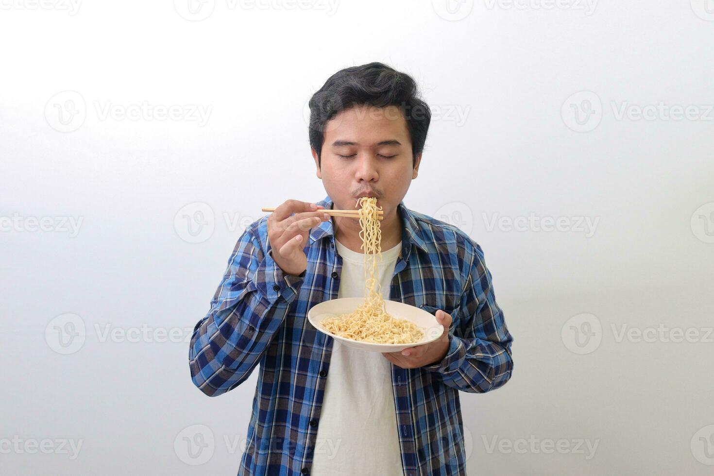 retrato de contento asiático hombre en azul tartán camisa comiendo delicioso instante tallarines con palillos servido en lámina. aislado imagen en blanco antecedentes foto