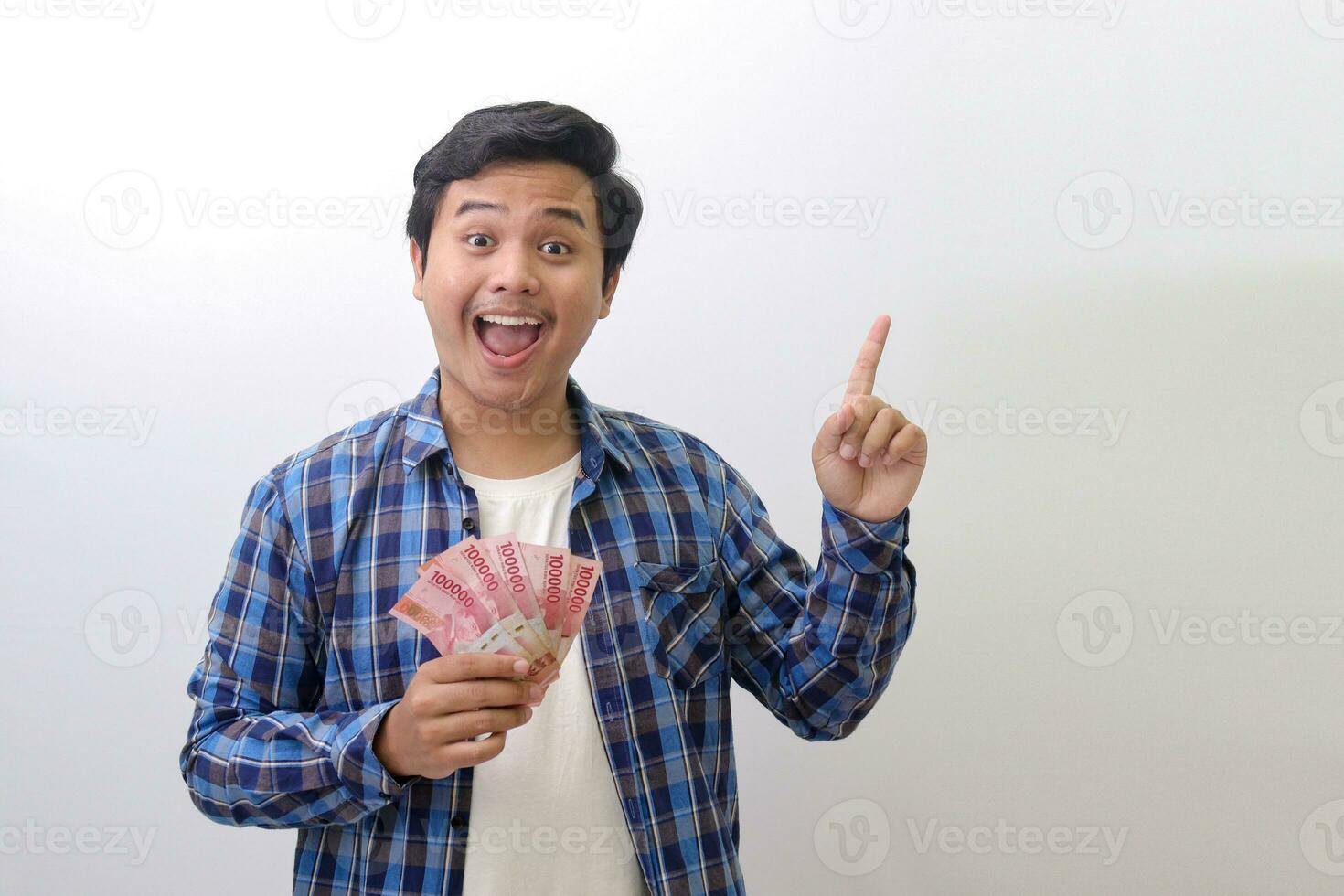 retrato de emocionado asiático hombre en azul tartán camisa en pie en contra blanco fondo, demostración uno cien mil rupia mientras señalando a el lado. financiero y ahorros concepto. foto