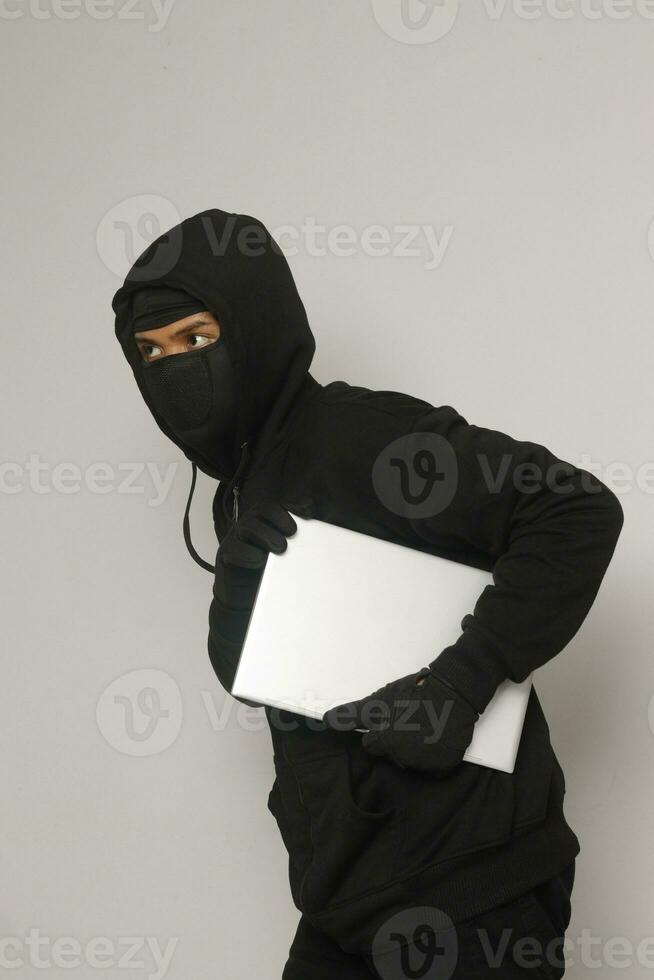 Mysterious robber thief man wearing black hoodie and mask stealing laptop and sneak out. Isolated image on gray background photo