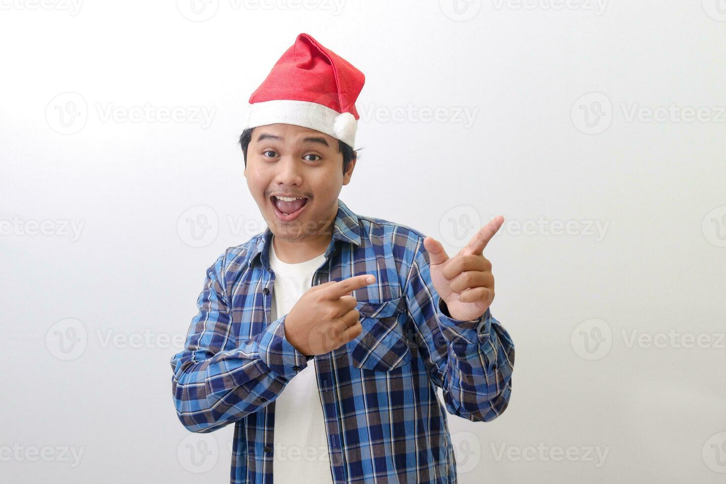 Portrait of excited Asian man in blue plaid shirt wearing santa hat smiling and looking at the camera pointing with two hands and fingers to the side. Isolated image on white background photo