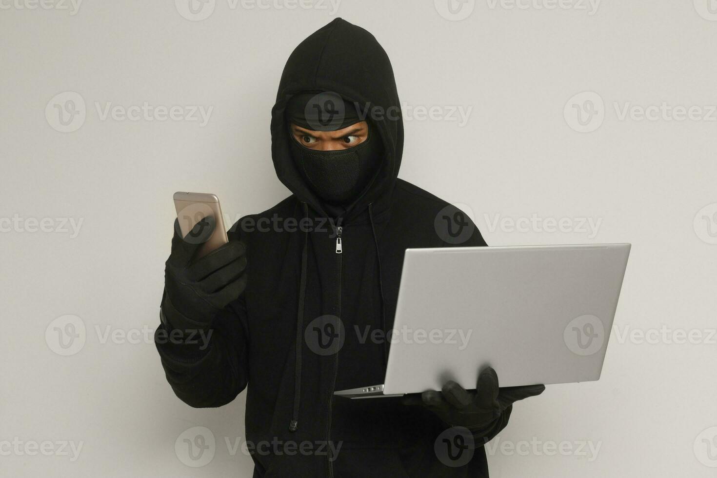 Portrait of mysterious man wearing black hoodie and mask doing hacking activity on laptop, hacker holding a mobile phone. Cyber security concept. Isolated image on gray background photo