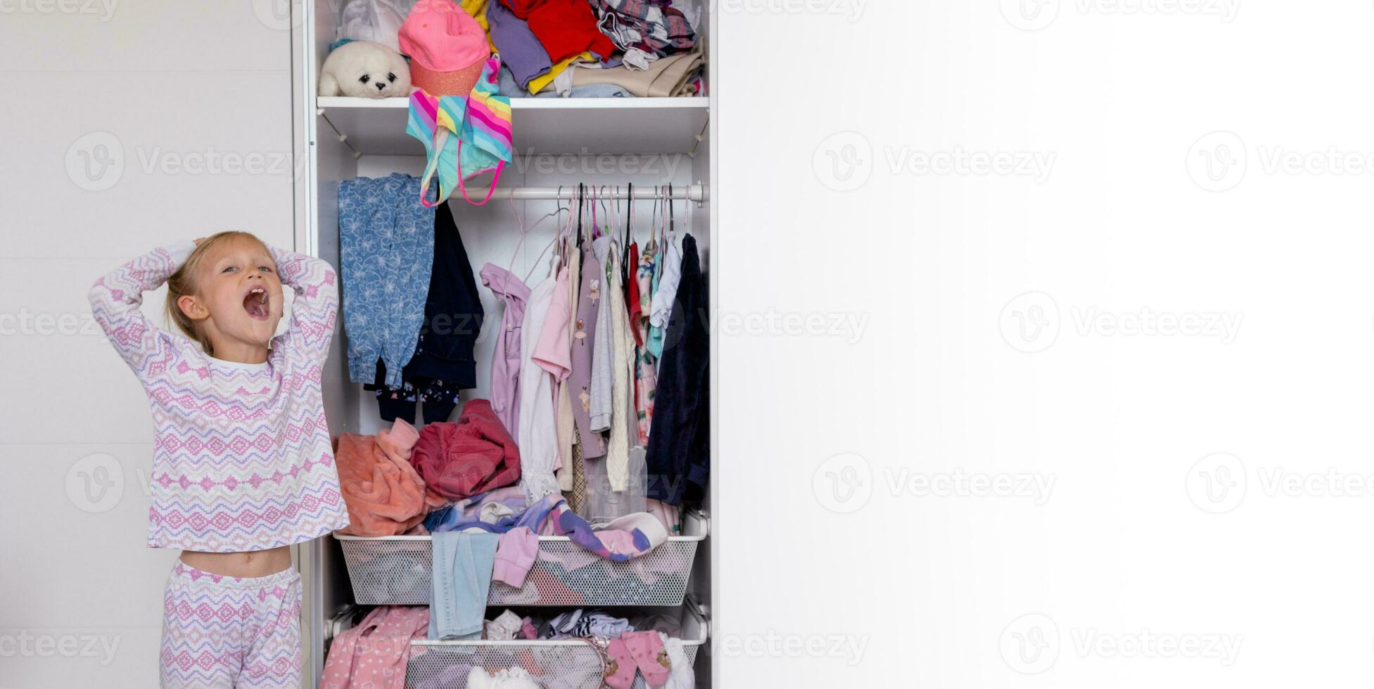 pequeño niña en rosado rosado pijama cuelga cosas en el armario, lío en el habitación foto