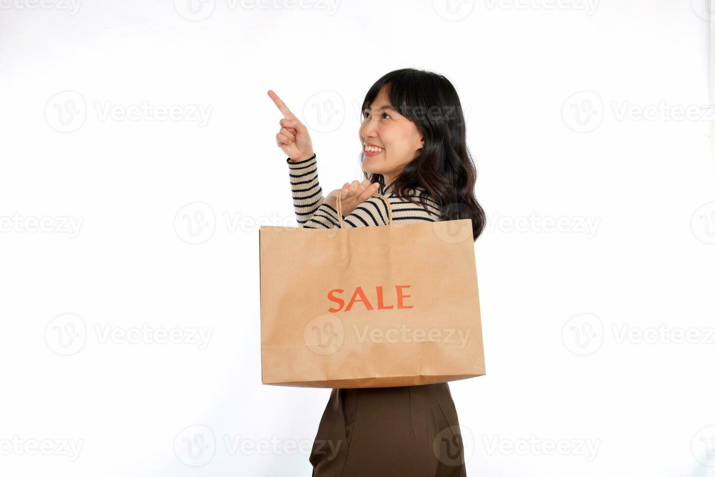 Beautiful young Asian woman on sweater cloth pointing up to copy space and looking at camera with smile face and sale shopping paper bag isolated on white background photo