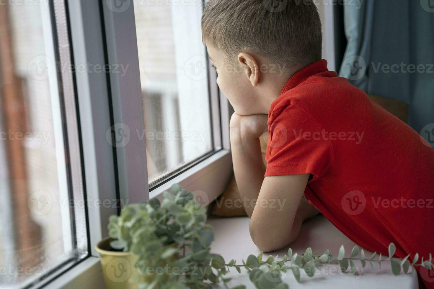 Adorable little blond kid boy sitting near window and looking on raindrops photo