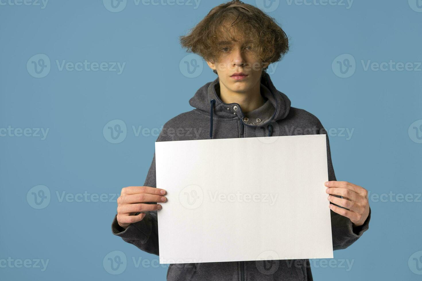 A serious, disgruntled teenager in a gray hoodie and wavy hair holds an empty poster on a blue background. photo