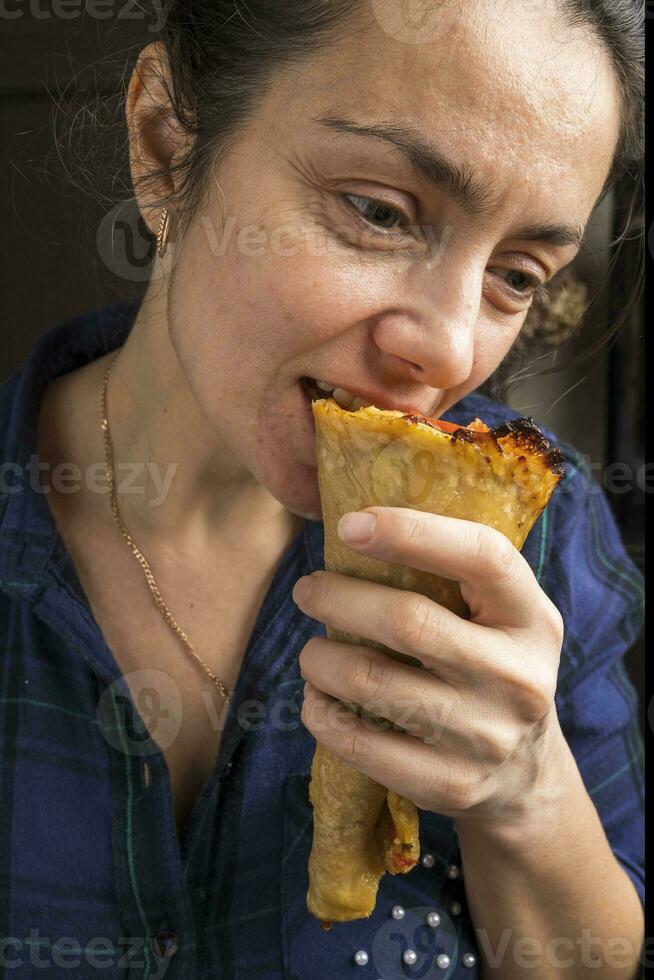 A pretty middle-aged woman in a purple plaid shirt is eating a pizza cone. Fast food. photo