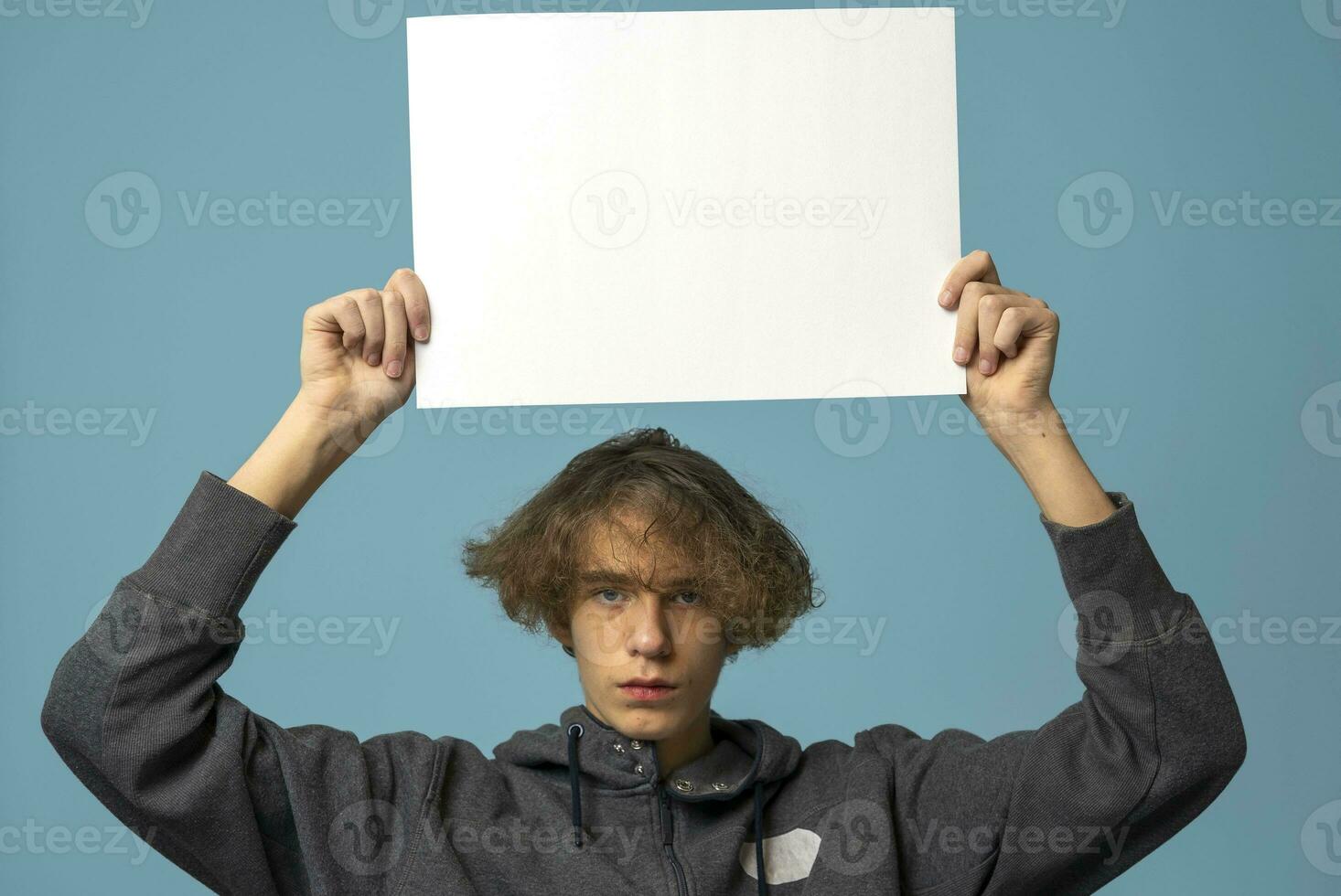 A serious, disgruntled teenager in a gray hoodie and wavy hair holds an empty poster on a blue background. photo