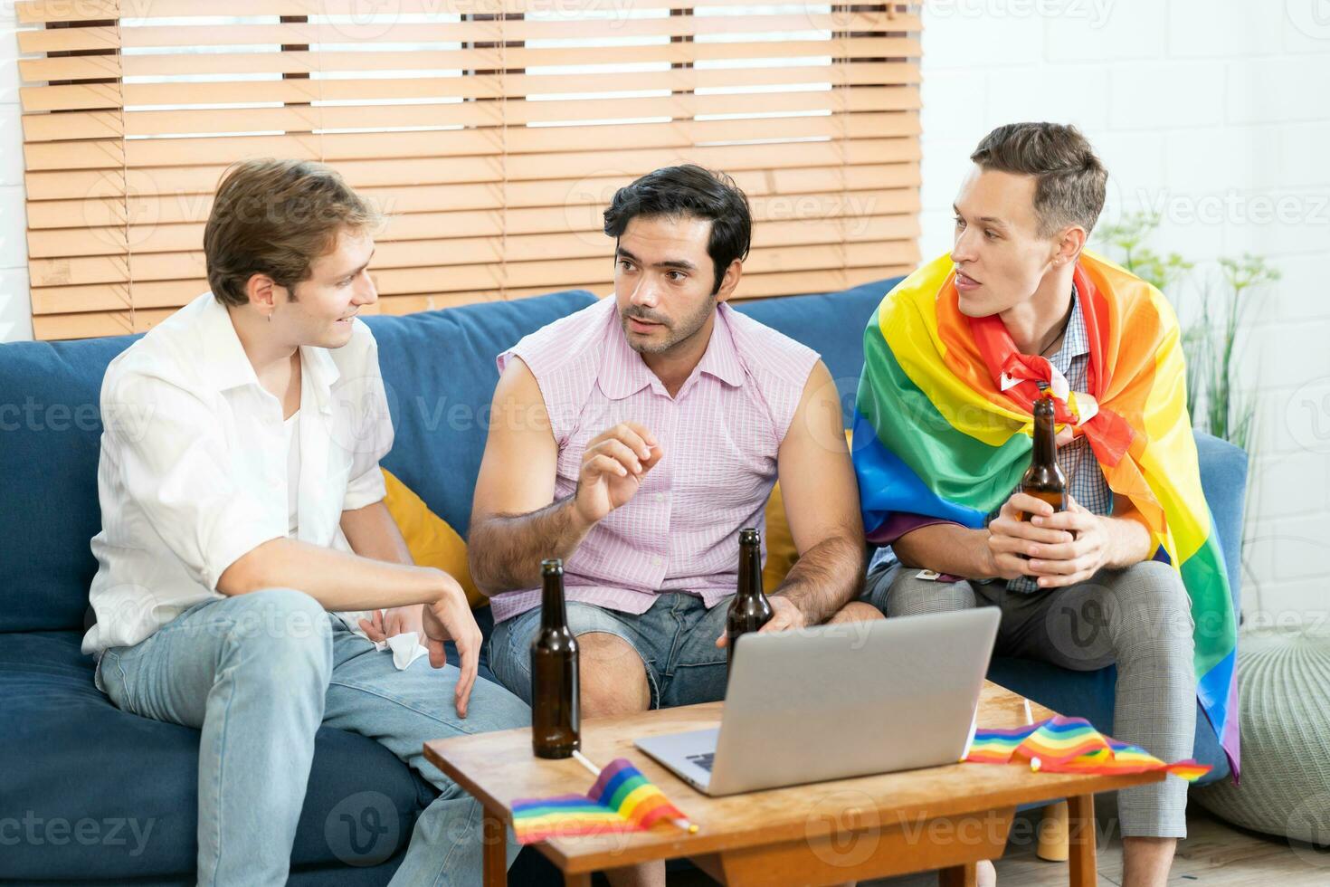 triple joven hombre sentado en el vivo habitación mientras utilizando ordenador portátil a vídeo llamada y participación y ondulación arco iris lgbt orgullo bandera. lgbtq personas estilo de vida. cerebrándose el orgullo día. foto