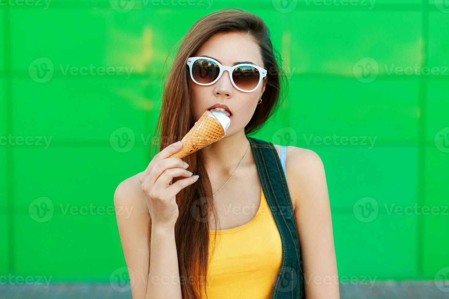 Beautiful young girl eating ice cream near the green wall.Stylish beautiful girl with ice-cream stands near green metal wall. photo