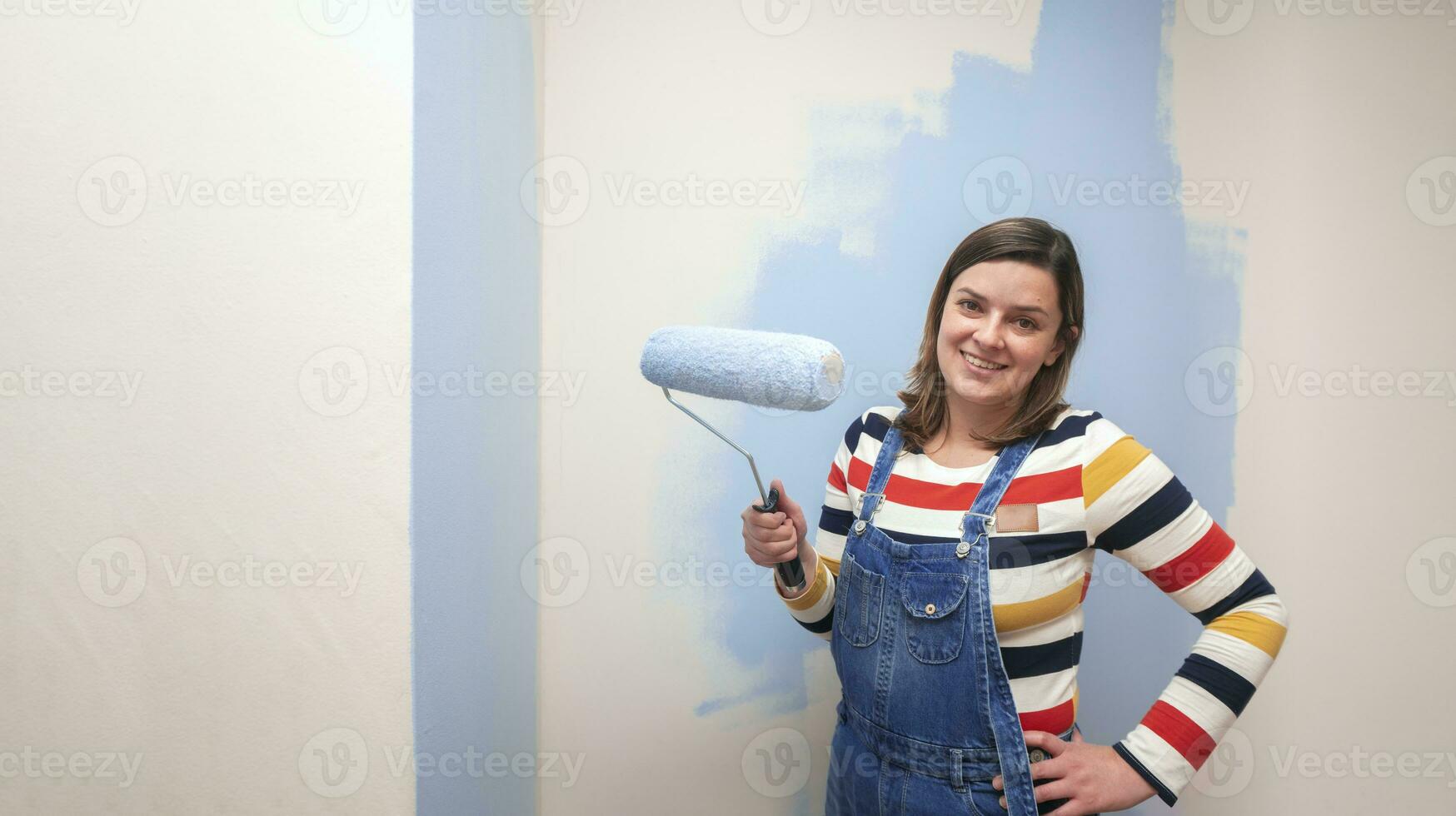 Beautiful woman dressed in overalls, smiling at camera with blue paint roller in her hand against half-painted white wall background photo