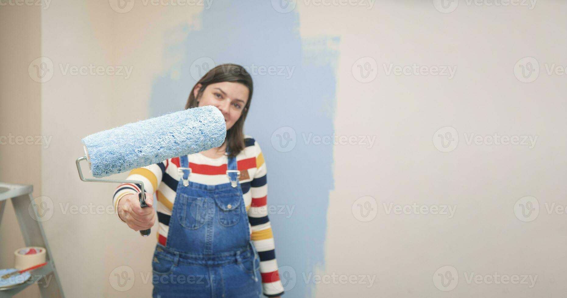roller with blue paint in the hand of a standing woman dressed in overalls and striped blouse, seen from the front, against background of half-painted white wall with strokes of blue paint photo
