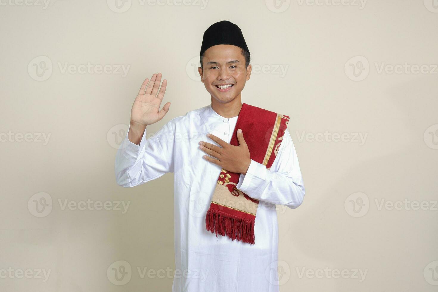 Portrait of attractive Asian muslim man in white shirt with skullcap swears with hands on chest and palms open, makes an oath of allegiance. Isolated image on blue background photo