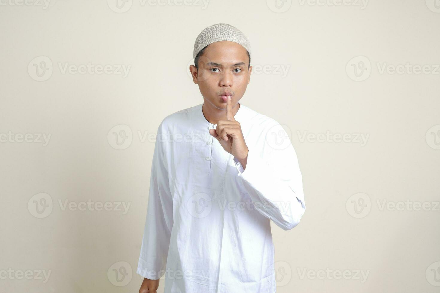 Portrait of attractive Asian muslim man in white shirt whispering malicious talk conversation, hand on mouth telling secret rumor. Isolated image on gray background photo