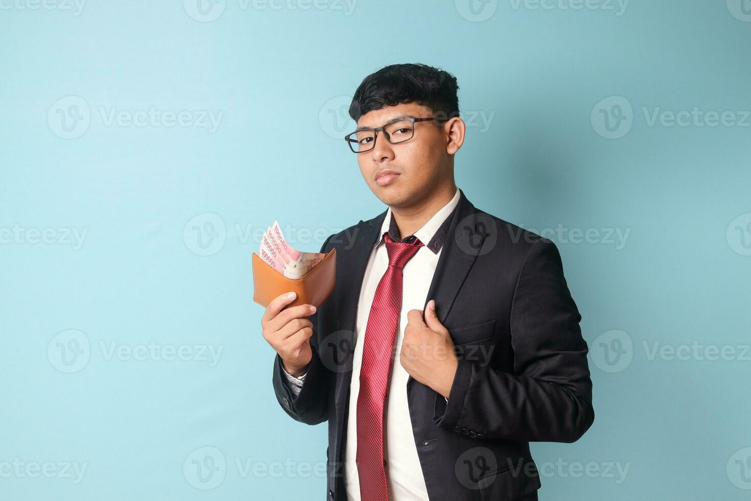 Portrait of young Asian business man in casual suit with confidence expressions fixing collar and holding leather wallet with thousand rupiahs. Isolated image on blue background photo